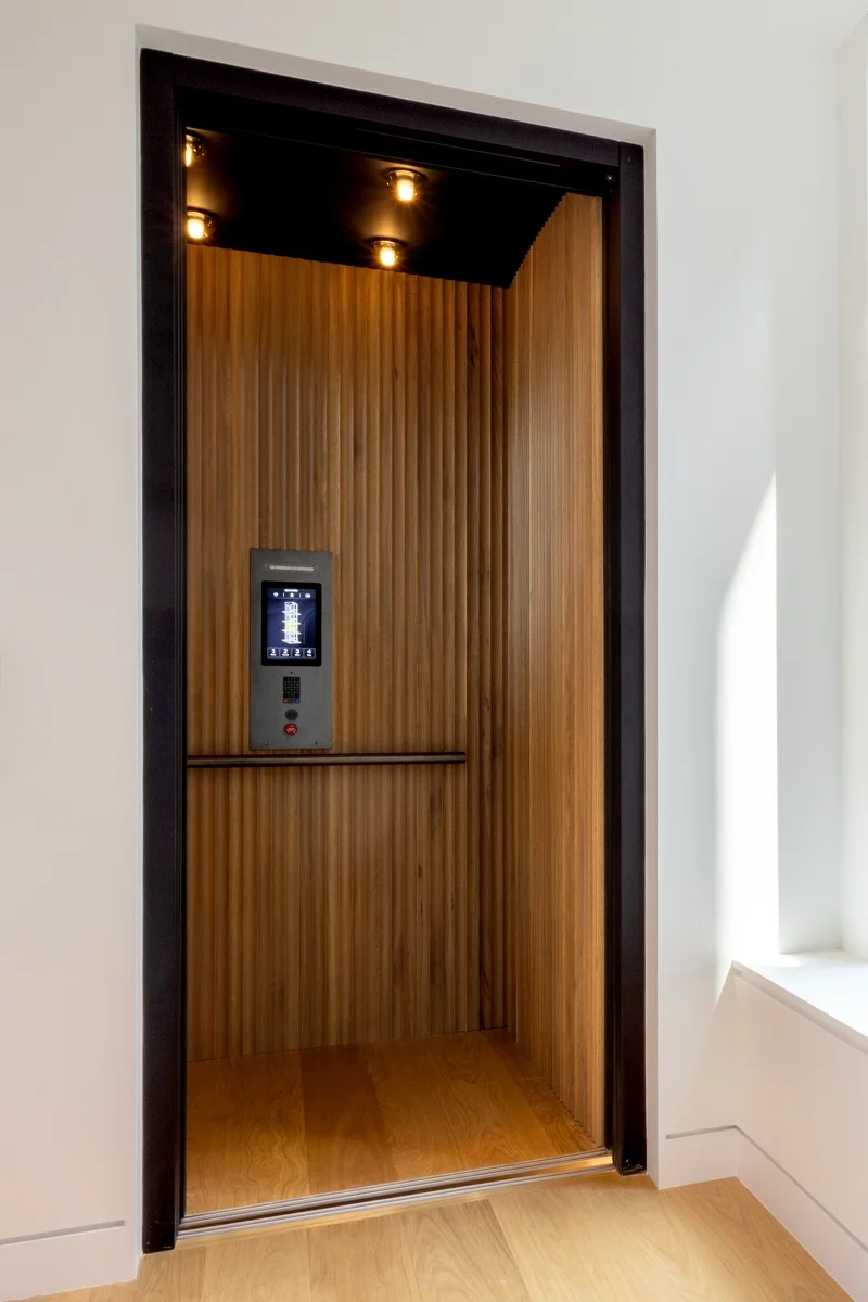 Home elevator interior featuring vertical wood‑slat wall panels, warm recessed ceiling lights, a touchscreen control panel on the side wall, and wood flooring that matches the surrounding room.
